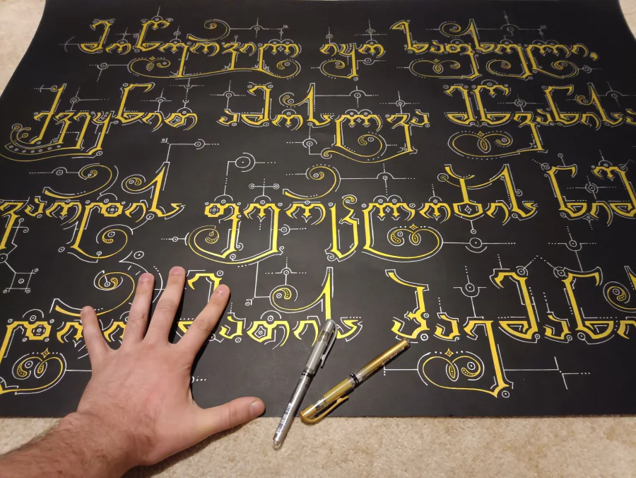 Text from The Knight in the Panther’s Skin in golden Georgian lettering on black paper, photographed in progress with a hand and two pens on it.