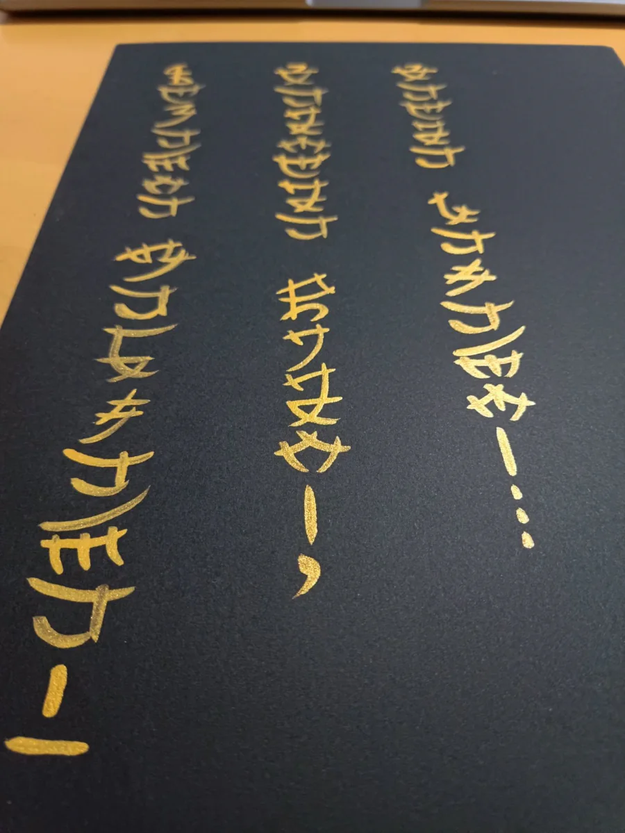 Three vertical lines of golden lettering on a black sheet, photographed at an angle on a wooden desk.
